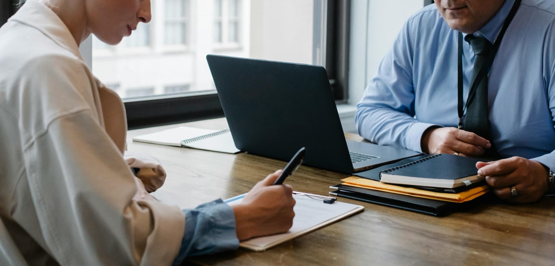 A woman and a man in a shirt and tie sit across from each other at a table. She is signing a bond. He is a financial advisor.